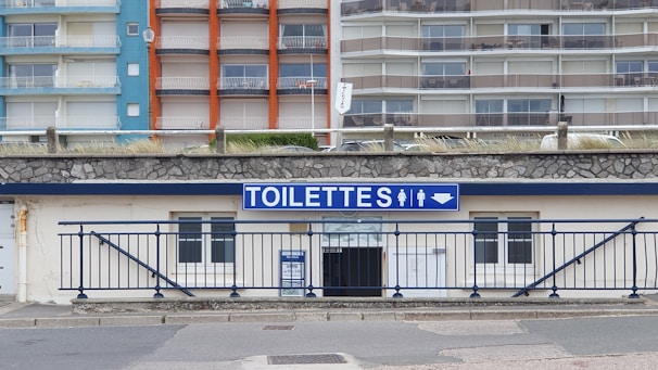 A public restroom facility with a prominent blue and white sign labeled 'TOILETTES' above the entrance. The building features a stone facade and is located at the base of a large apartment complex. The apartments have colorful facades with blue, orange, and beige sections.
