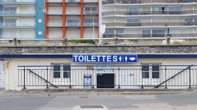 A public restroom facility with a prominent blue and white sign labeled 'TOILETTES' above the entrance. The building features a stone facade and is located at the base of a large apartment complex. The apartments have colorful facades with blue, orange, and beige sections.