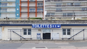 A public restroom facility with a prominent blue and white sign labeled 'TOILETTES' above the entrance. The building features a stone facade and is located at the base of a large apartment complex. The apartments have colorful facades with blue, orange, and beige sections.