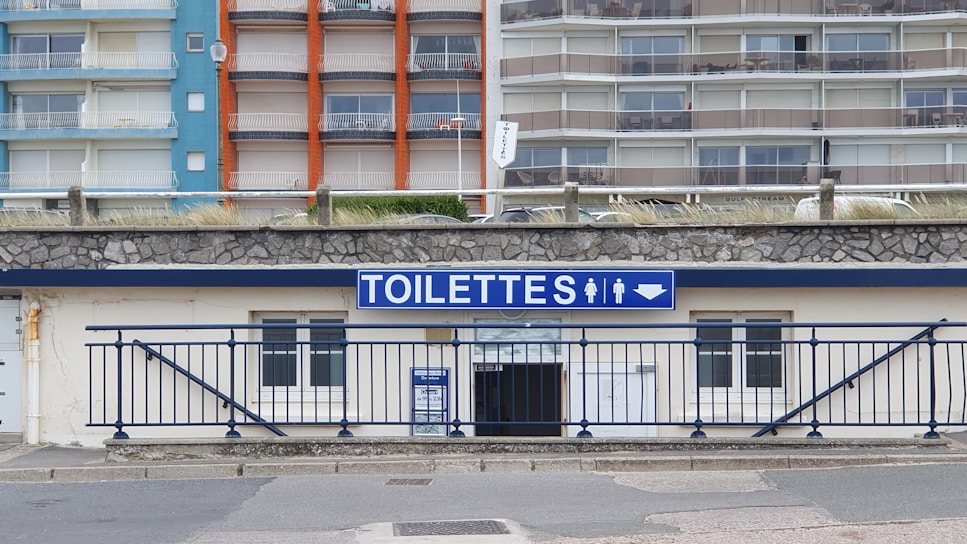 A public restroom facility with a prominent blue and white sign labeled 'TOILETTES' above the entrance. The building features a stone facade and is located at the base of a large apartment complex. The apartments have colorful facades with blue, orange, and beige sections.