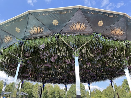 A decorative structure featuring a canopy with ornate designs and floral decorations. The ceiling is adorned with golden floral patterns against a dark background, and the underside is covered with lush greenery and hanging purple and blue flowers. The frame is supported by mirrored pillars beneath a clear blue sky.