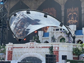 A theatrical stage set resembling a castle, featuring medieval architectural elements and decorative banners. The castle structure has stone textures and red accents, with greenery and flowers adorning parts of the set. A large, translucent canopy supported by a metal framework is overhead, lit with various colored lights.