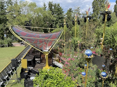 A beautifully arranged outdoor stage set up amidst lush greenery and vibrant flowers. There is a large, intricately designed canopy with red and gray patterns supported by golden structures. Surrounding the stage are tall poles with decorative lighting fixtures shaped like flames and reflective blue spheres. Various stage equipment and speakers are positioned strategically, creating an organized setup for a performance or event.