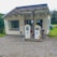 A small, vintage gas station with two old BP fuel pumps positioned in front of a simple, single-story building. The building has a slanted roof and large windows, surrounded by a rural landscape with greenery and trees in the background.