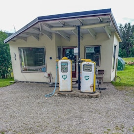 A small, vintage gas station with two old BP fuel pumps positioned in front of a simple, single-story building. The building has a slanted roof and large windows, surrounded by a rural landscape with greenery and trees in the background.