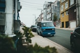 A blue van is parked on a sloping urban street lined with houses. The buildings vary in color and have multiple stories. There are also some plants and blurred foliage in the foreground, with visible street lights and utility poles and wires overhead.