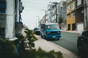 A blue van is parked on a sloping urban street lined with houses. The buildings vary in color and have multiple stories. There are also some plants and blurred foliage in the foreground, with visible street lights and utility poles and wires overhead.