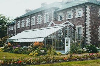 A beautiful stone building with a green metal roof is complemented by an adjoining glass greenhouse. The greenhouse is surrounded by vibrant and diverse flower beds, creating a lush garden setting. The combination of brickwork and glass accentuates both man-made and natural elements. Soft natural light adds a serene and fresh ambiance to the scene.