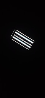 A rectangular fluorescent ceiling light fixture, glowing brightly against a dark background. The light consists of four parallel tubes, emitting a white glow that contrasts with the surrounding darkness.