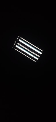 A rectangular fluorescent ceiling light fixture, glowing brightly against a dark background. The light consists of four parallel tubes, emitting a white glow that contrasts with the surrounding darkness.