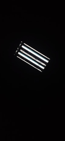 A rectangular fluorescent ceiling light fixture, glowing brightly against a dark background. The light consists of four parallel tubes, emitting a white glow that contrasts with the surrounding darkness.