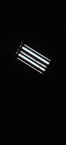 A rectangular fluorescent ceiling light fixture, glowing brightly against a dark background. The light consists of four parallel tubes, emitting a white glow that contrasts with the surrounding darkness.