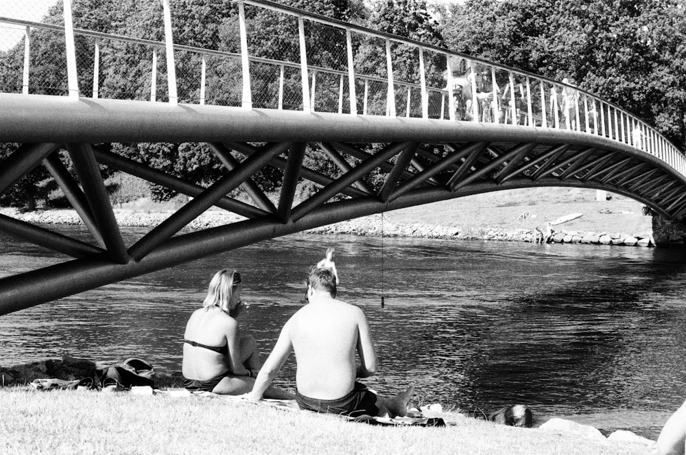 a man and a woman sitting on the bank of a river under a bridge