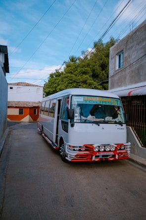 A white bus with red accents is parked on a narrow street lined with buildings and greenery. Overhead, a network of electrical wires is visible against a backdrop of a bright blue sky.
