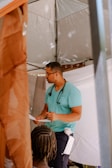 A person wearing a teal shirt and dark pants stands under a tent. They have a stethoscope around their neck and are holding a piece of paper, appearing to be in conversation with another individual who is partially visible with braided hair.