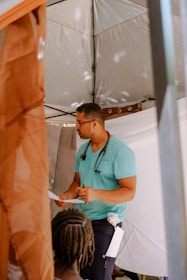 A person wearing a teal shirt and dark pants stands under a tent. They have a stethoscope around their neck and are holding a piece of paper, appearing to be in conversation with another individual who is partially visible with braided hair.