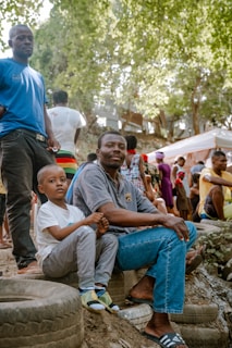 Several people are gathered outdoors in a wooded area with large trees and dappled sunlight filtering through the leaves. A man wearing a gray shirt and blue jeans sits on a stack of tires, holding a young boy with a serious expression. Nearby, other people are standing or sitting, engaged in conversation or watching something off-camera. Tents or temporary structures in the background suggest a casual or community event.