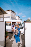 A small group of people are boarding a white bus parked on the side of a street next to several multi-story buildings. The individuals are carrying backpacks and casual attire, indicating they might be travelers or commuters. The sky is clear and blue, suggesting a sunny day.