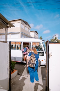A small group of people are boarding a white bus parked on the side of a street next to several multi-story buildings. The individuals are carrying backpacks and casual attire, indicating they might be travelers or commuters. The sky is clear and blue, suggesting a sunny day.