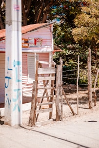 A small wooden house with pink siding and a rusted metal roof stands next to a rudimentary wooden fence. A makeshift wooden ladder leans against the fence. There is graffiti on a nearby utility pole, and a mix of trees and greenery partially shade the scene.