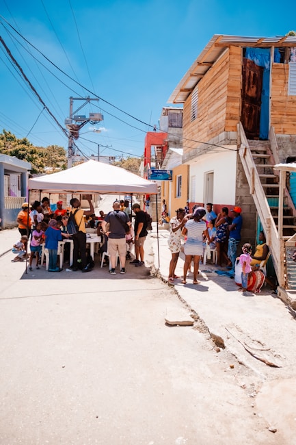 A group of people gathered around a table covered by a canopy situated in an urban neighborhood setting. They are engaged in conversation or community activities. The area has various houses and buildings with visible electric poles and wires overhead. Children and adults are present, some standing and others seated.