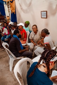 A group of people are seated in a room, engaging in conversation. Several individuals, including both men and women, are sitting on plastic chairs. One woman appears to be a health worker, identifiable by a stethoscope around her neck. Another person appears to be speaking directly to her. The room is decorated with blue and yellow drapes, and a framed picture or sign labeled 'CLASSE DEBORA' is visible on the wall.