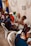 A group of people are seated in a room, engaging in conversation. Several individuals, including both men and women, are sitting on plastic chairs. One woman appears to be a health worker, identifiable by a stethoscope around her neck. Another person appears to be speaking directly to her. The room is decorated with blue and yellow drapes, and a framed picture or sign labeled 'CLASSE DEBORA' is visible on the wall.