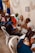 A group of people are seated in a room, engaging in conversation. Several individuals, including both men and women, are sitting on plastic chairs. One woman appears to be a health worker, identifiable by a stethoscope around her neck. Another person appears to be speaking directly to her. The room is decorated with blue and yellow drapes, and a framed picture or sign labeled 'CLASSE DEBORA' is visible on the wall.