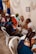 A group of people are seated in a room, engaging in conversation. Several individuals, including both men and women, are sitting on plastic chairs. One woman appears to be a health worker, identifiable by a stethoscope around her neck. Another person appears to be speaking directly to her. The room is decorated with blue and yellow drapes, and a framed picture or sign labeled 'CLASSE DEBORA' is visible on the wall.