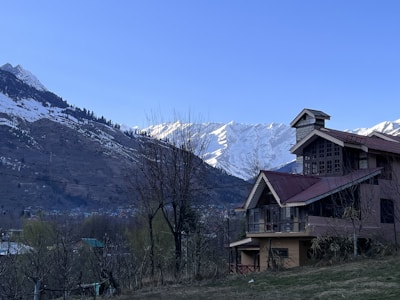 A rustic house with red roofs is situated in a mountainous area, surrounded by leafless trees and grass. In the background, there are snow-capped mountains under a clear blue sky.