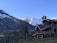 A rustic house with red roofs is situated in a mountainous area, surrounded by leafless trees and grass. In the background, there are snow-capped mountains under a clear blue sky.