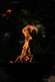 Flames rise from a black grill, set against a dark, blurry background. The fire twists and dances upwards with bright orange and yellow hues, giving a vivid contrast to the surrounding darkness.