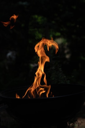 Flames rise from a black grill, set against a dark, blurry background. The fire twists and dances upwards with bright orange and yellow hues, giving a vivid contrast to the surrounding darkness.