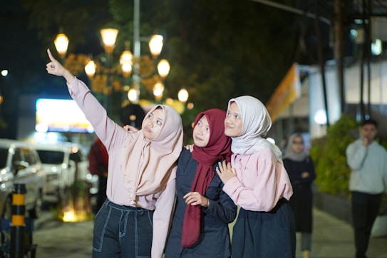 Three women wearing hijabs stand on a well-lit street at night. The woman on the left is pointing upwards, possibly at something in the sky. They are surrounded by street lamps, a parked car, and blurred people in the background.