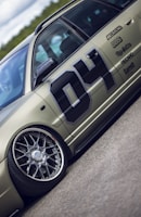 A detailed view of a customized car with prominent wheel rims and a matte finish. The car features decals and sponsor logos, highlighting brands such as BBS and Recaro, with a large number 04 on the side. The vehicle is positioned on a smooth road surface, with greenery visible in the background under a partly cloudy sky.