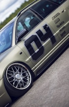A detailed view of a customized car with prominent wheel rims and a matte finish. The car features decals and sponsor logos, highlighting brands such as BBS and Recaro, with a large number 04 on the side. The vehicle is positioned on a smooth road surface, with greenery visible in the background under a partly cloudy sky.