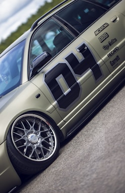 A detailed view of a customized car with prominent wheel rims and a matte finish. The car features decals and sponsor logos, highlighting brands such as BBS and Recaro, with a large number 04 on the side. The vehicle is positioned on a smooth road surface, with greenery visible in the background under a partly cloudy sky.