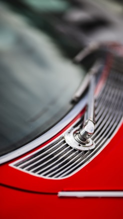Close-up view of a vintage car's windshield wiper mechanism and chrome detailing. The red body of the car adds a vibrant contrast to the metal elements and the slightly blurred background.