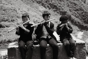 Three children sit on a stone ledge playing flutes. They are dressed in dark clothing, and their expressions suggest concentration and absorption in the music. The background features a rugged landscape with hills and vegetation.