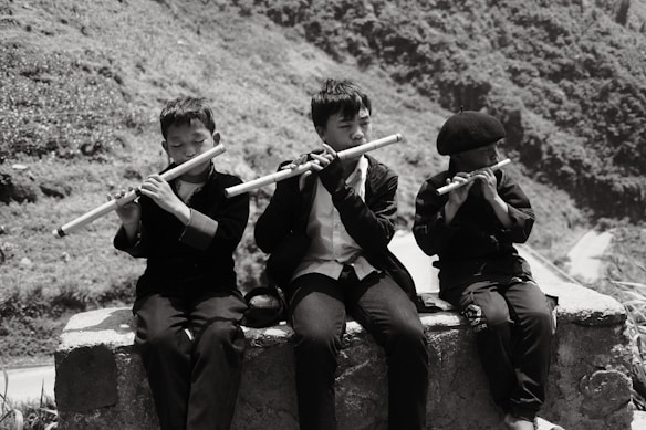 Three children sit on a stone ledge playing flutes. They are dressed in dark clothing, and their expressions suggest concentration and absorption in the music. The background features a rugged landscape with hills and vegetation.