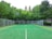 A tennis court surrounded by a metal fence, situated in a lush green area with various trees. The court has a clean, green surface marked with white lines, and there is a pole with lights on the left side. The background is filled with dense foliage, adding a serene and natural ambiance.