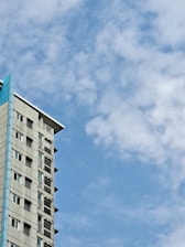 A multi-story building with a modern design, featuring large windows and a blue accent on its corner. The sky is filled with scattered fluffy clouds against a bright blue background, creating a peaceful atmosphere.