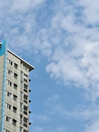 A multi-story building with a modern design, featuring large windows and a blue accent on its corner. The sky is filled with scattered fluffy clouds against a bright blue background, creating a peaceful atmosphere.