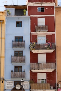 Two adjacent residential buildings feature distinct colors and styles. The left building is light blue with several wooden balconies, while the right building is red with white trim and also features balconies adorned with colorful flowers. Both buildings have closed shutters on some windows. At the ground level, there are signs and a clock between the buildings advertising the sale of various items like coins and stamps.