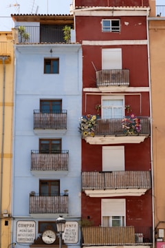 Two adjacent residential buildings feature distinct colors and styles. The left building is light blue with several wooden balconies, while the right building is red with white trim and also features balconies adorned with colorful flowers. Both buildings have closed shutters on some windows. At the ground level, there are signs and a clock between the buildings advertising the sale of various items like coins and stamps.