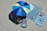 A large blue and green beach umbrella casts shade over two striped beach chairs on a sandy beach. Next to the chairs, a large blue towel covers part of the sand with smaller items like a can and an extra towel nearby. A striped blue beach mat sits to the right, along with a patterned bag.