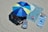 A large blue and green beach umbrella casts shade over two striped beach chairs on a sandy beach. Next to the chairs, a large blue towel covers part of the sand with smaller items like a can and an extra towel nearby. A striped blue beach mat sits to the right, along with a patterned bag.