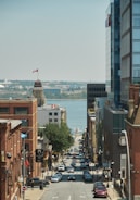 A busy city street leading down towards a waterfront. Tall buildings line both sides of the street, and a Canadian flag is visible atop a building dome on the left. Numerous cars are parked along the street and driving along it. Trees can be seen along the street, and the sky is clear and blue with distant land visible across the water.
