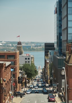 A busy city street leading down towards a waterfront. Tall buildings line both sides of the street, and a Canadian flag is visible atop a building dome on the left. Numerous cars are parked along the street and driving along it. Trees can be seen along the street, and the sky is clear and blue with distant land visible across the water.