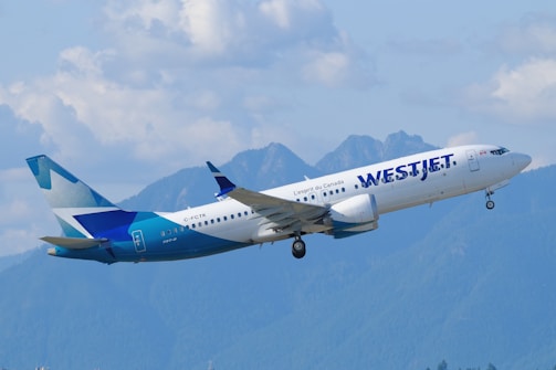 A passenger airplane with the WestJet logo is flying in the sky. The backdrop includes a range of blue mountains and a partly cloudy sky.